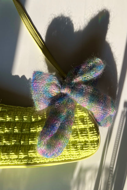 Lake Kingdom - Starpot Studio. Blue, pink, green and yellow tie-dye knitted bow resting atop a neon green ruched shoulder bag on a white background.