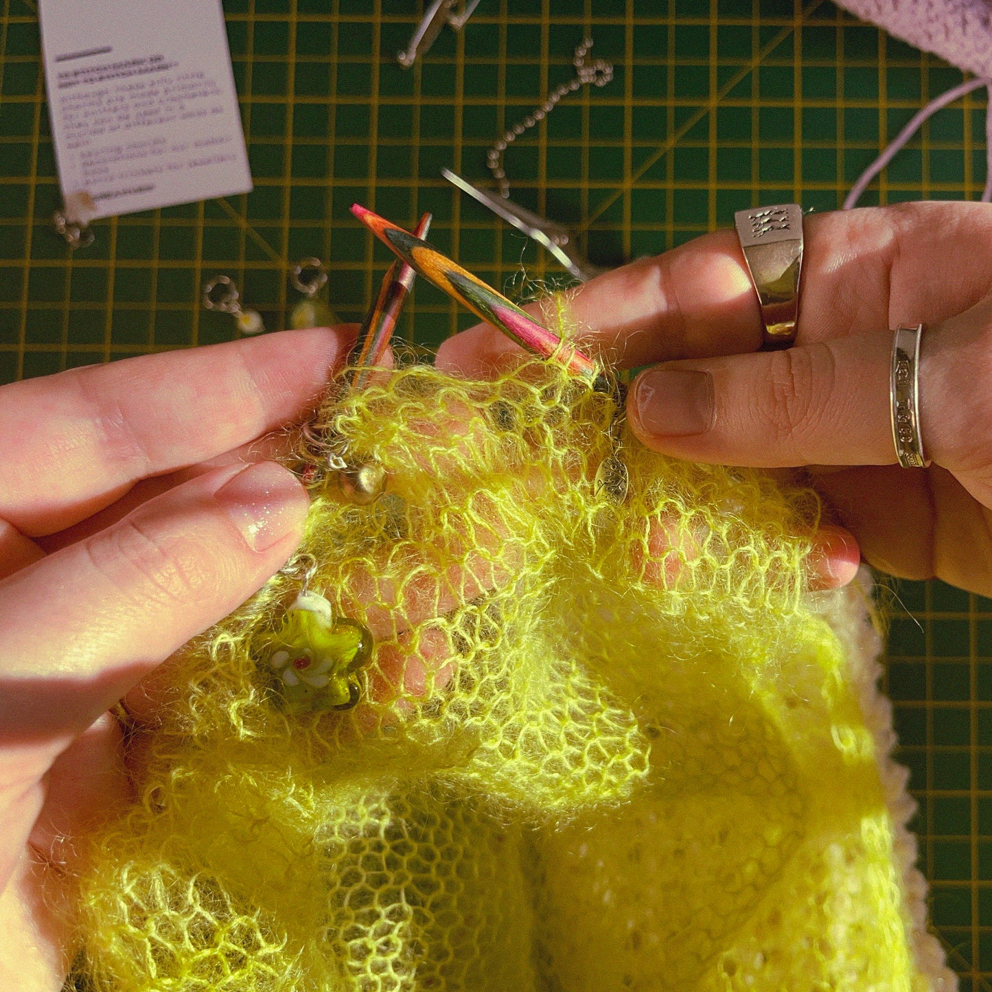 3 ring stitch markers for knitting being used on a neon green knit mohair piece. One stitch marker has a green lampwork flower bead on it, another has a metal oval bead with a flower pattern engraved into it on it and the third has a small green dyed freshwater pearl on it.