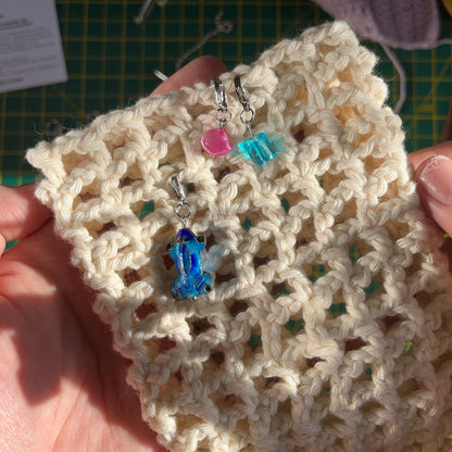 Three clasp stitch markers for crochet being used on a cream crocheted mesh piece. One stitch marker has a pink dyed freshwater pearl on, another has a bright blue cylindrical glass bead on and the third has a blue enamel and brass frog bead on it.