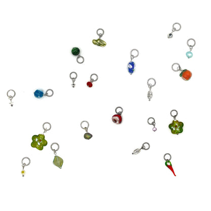 A large selection of ring stitch markers for knitting scattered across a white background. Each stitch marker has a different charm hanging from it: some are freshwater pearls, others are metal alloy charms and there are a few which have glass lampwork beads hanging from them. 