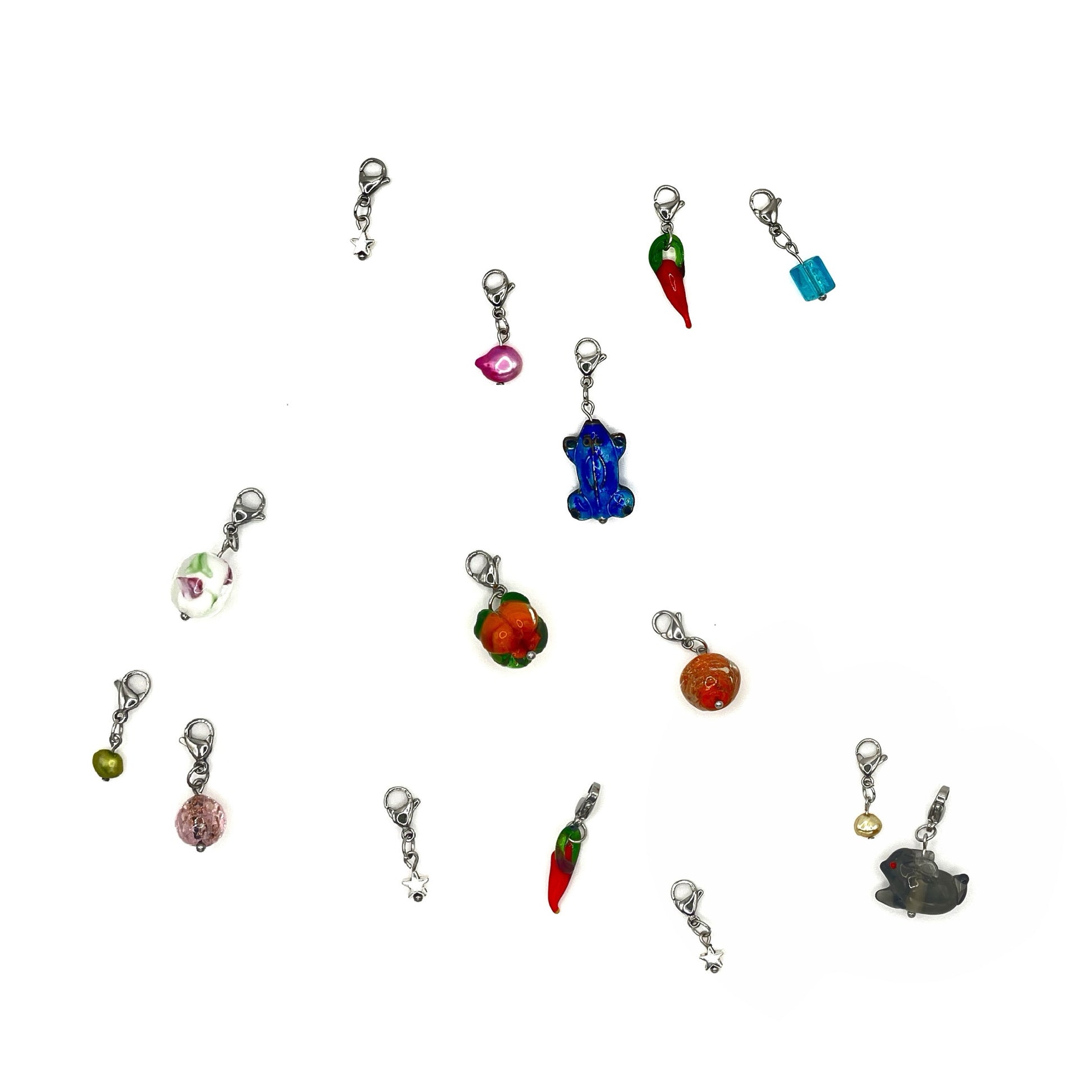 A selection of clasp stitch markers for crochet scattered across a white background. Each stitch marker has a different charm hanging from it: some are freshwater pearls, others are metal alloy charms and there are a few which have glass lampwork beads hanging from them.